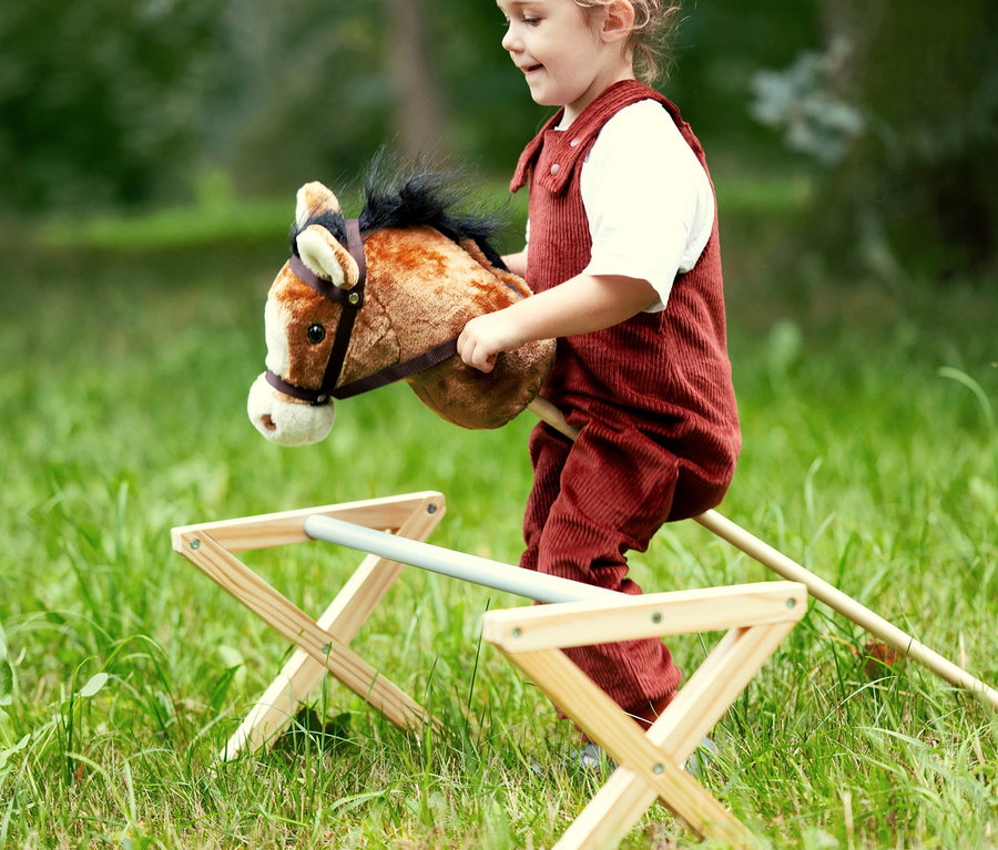 Mädchen in rotem Overall reitet auf einem braunen small foot Steckenpferd »Nico« auf einer Wiese.