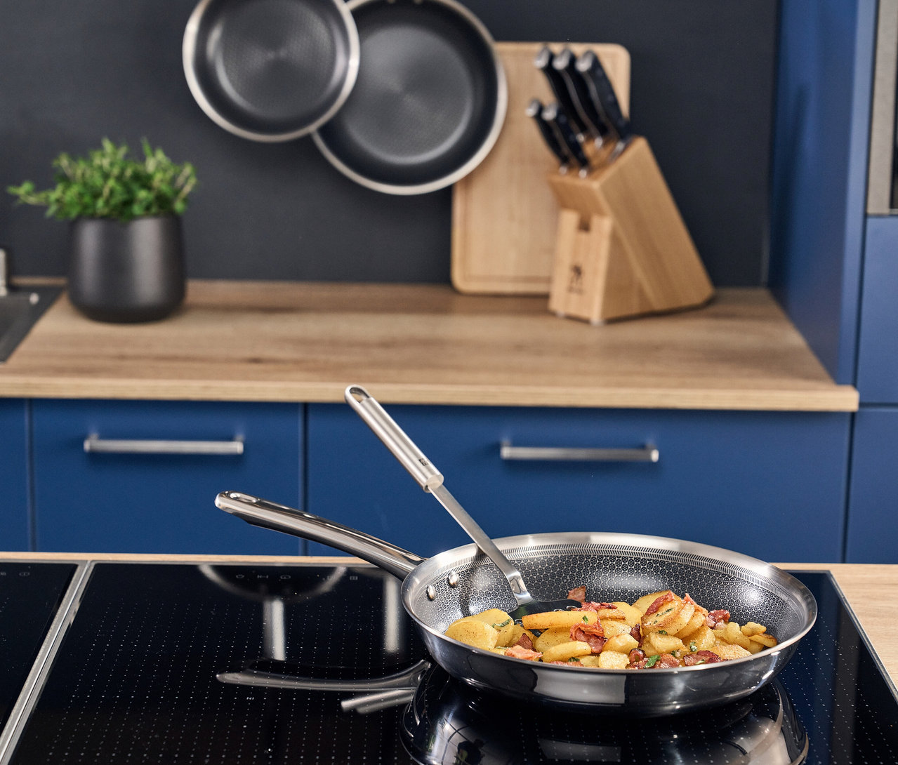 Eine Henckels by ZWILLING »Valley« Bratpfanne mit Wabenmuster, gefüllt mit Bratkartoffeln und Speck, steht auf einem Induktionskochfeld. Ein Metallwender ist in der Pfanne. Im Hintergrund hängen zwei weitere Pfannen und ein Messerblock steht auf der Holzarbeitsplatte einer blauen Küche.