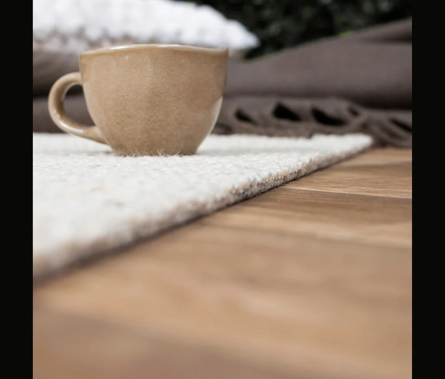 Eine beige Keramiktasse steht auf einem hellen, strukturierten Teppich auf einem Holzboden. Im Hintergrund sind eine braune Decke und Grünpflanzen unscharf zu sehen.