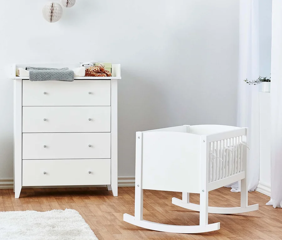 Ein Zimmer mit einer weißen Kommode, auf der ein graues Handtuch und Kinderkleidung liegen, und einer weißen Hoppekids Babywiege, ca. 40 x 80 cm.
