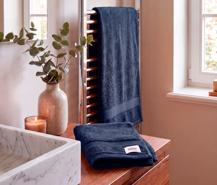 Zwei blaue Handtücher sind in einem Badezimmer ausgestellt. Ein blaues Handtuch der SCHÖNER WOHNEN-Kollektion liegt gefaltet auf einer Holzablage neben einem Marmorwaschbecken, einer brennenden Kerze und einer Vase mit Eukalyptuszweigen. Ein weiteres blaues Handtuch hängt im Hintergrund an einem verchromten Handtuchwärmer, nahe einem Fenster.