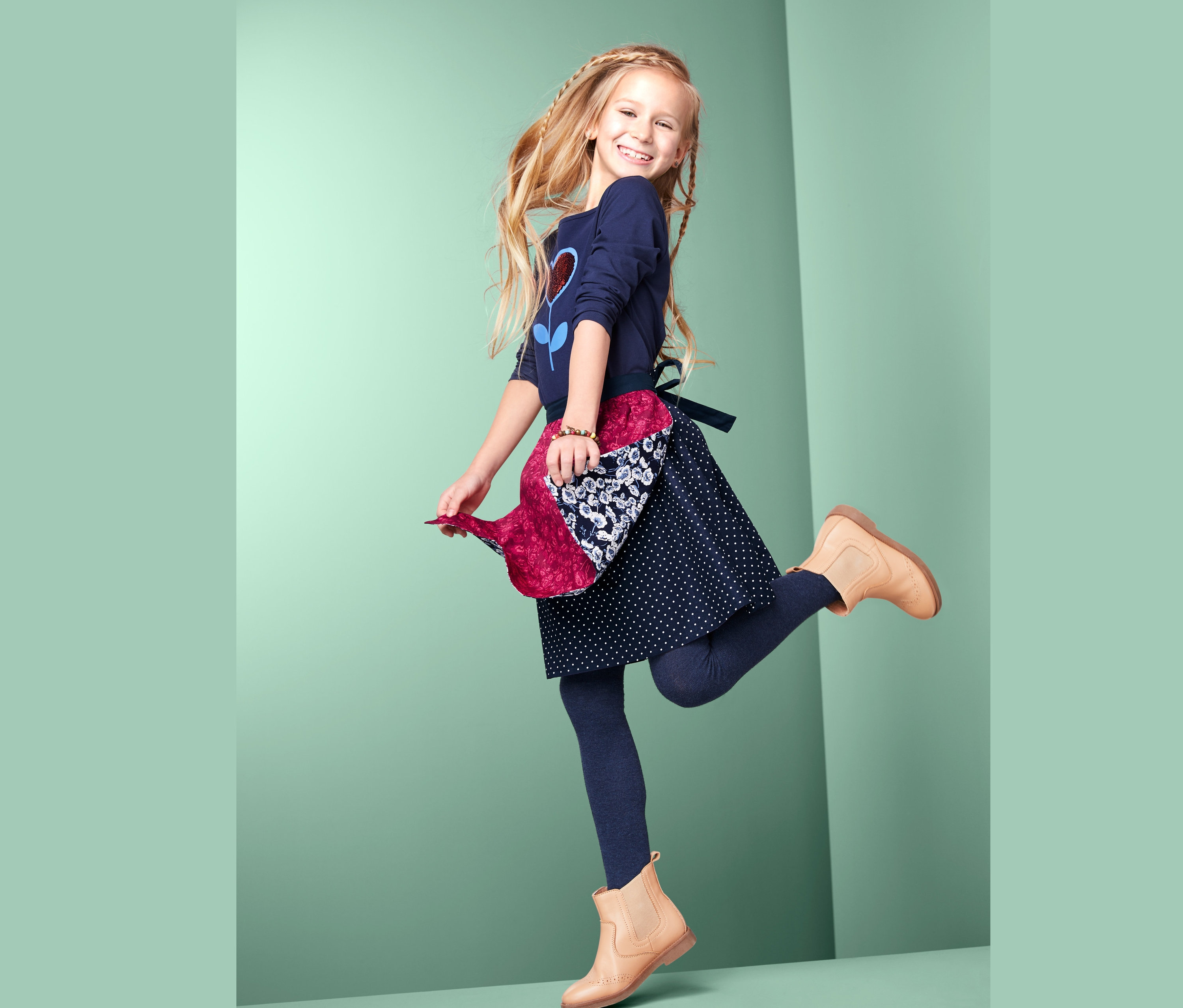 Lächelndes Mädchen hüpft vor hellgrünem Hintergrund in Rock mit Wendeschürze, blauen Leggings und braunen Stiefeln.