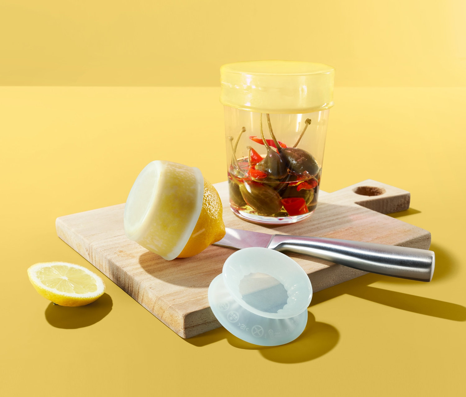 Auf gelbem Grund liegen ein Schneidebrett mit einem Glas Chilischoten, einer Zitrone und 3 Silikon-Frischhaltedeckeln.