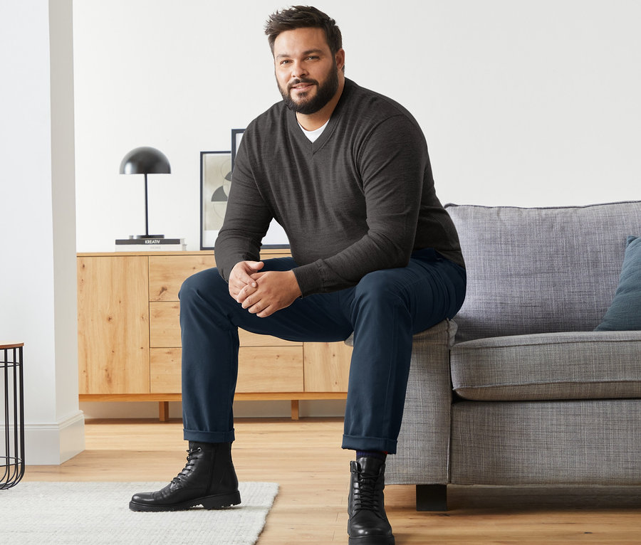 Ein Mann sitzt auf einem Sofa und trägt einen anthrazitfarbenen Merino-Pullover mit V-Ausschnitt, blaue Chinos und Leder-Boots.