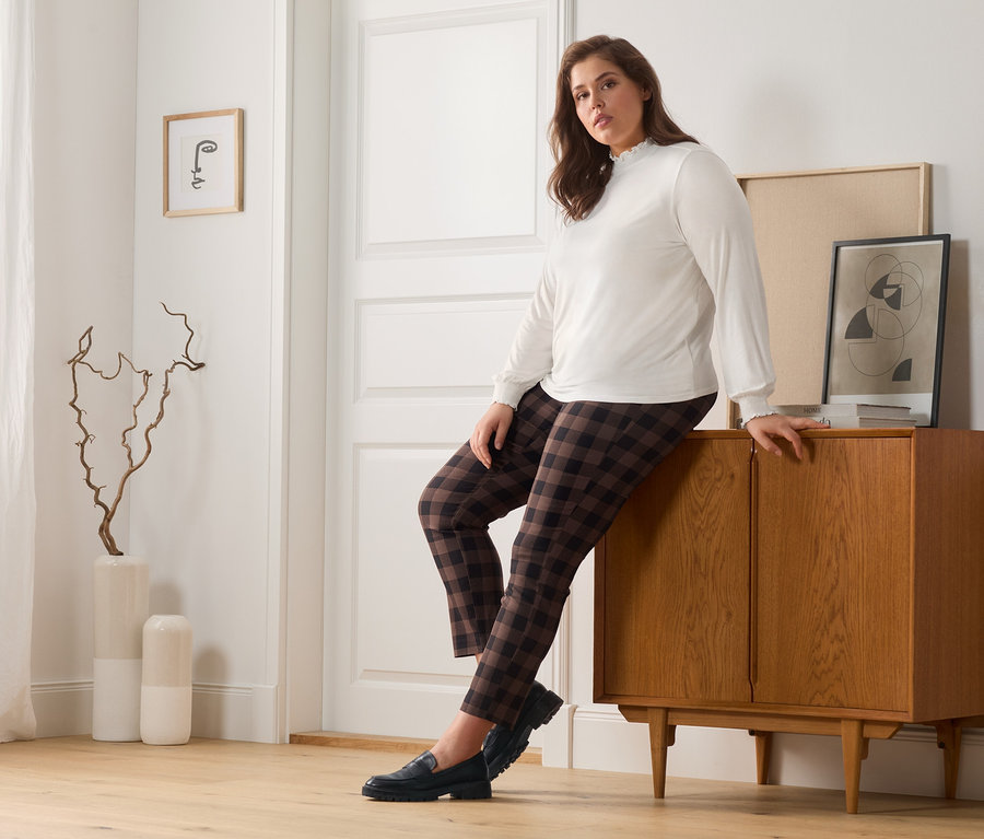 Frau lehnt an einer Holzkommode, trägt eine karierte Stretchhose, ein Blusenshirt mit Stehkragen und Leder-Loafer.