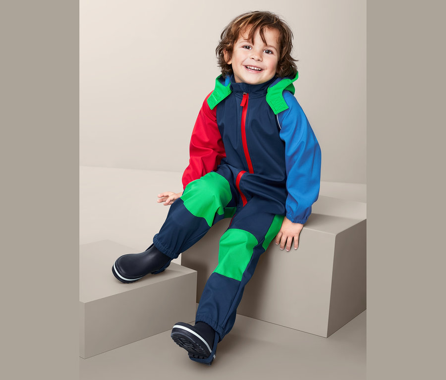 Ein lächelnder Junge sitzt und trägt einen Softshell-Einteiler aus recyceltem Material und Gummistiefel.