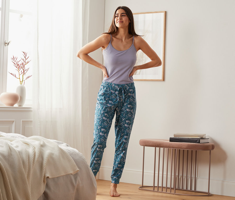 Eine Frau steht im Schlafzimmer und trägt eine Jersey-Relaxhose mit Alloverprint.