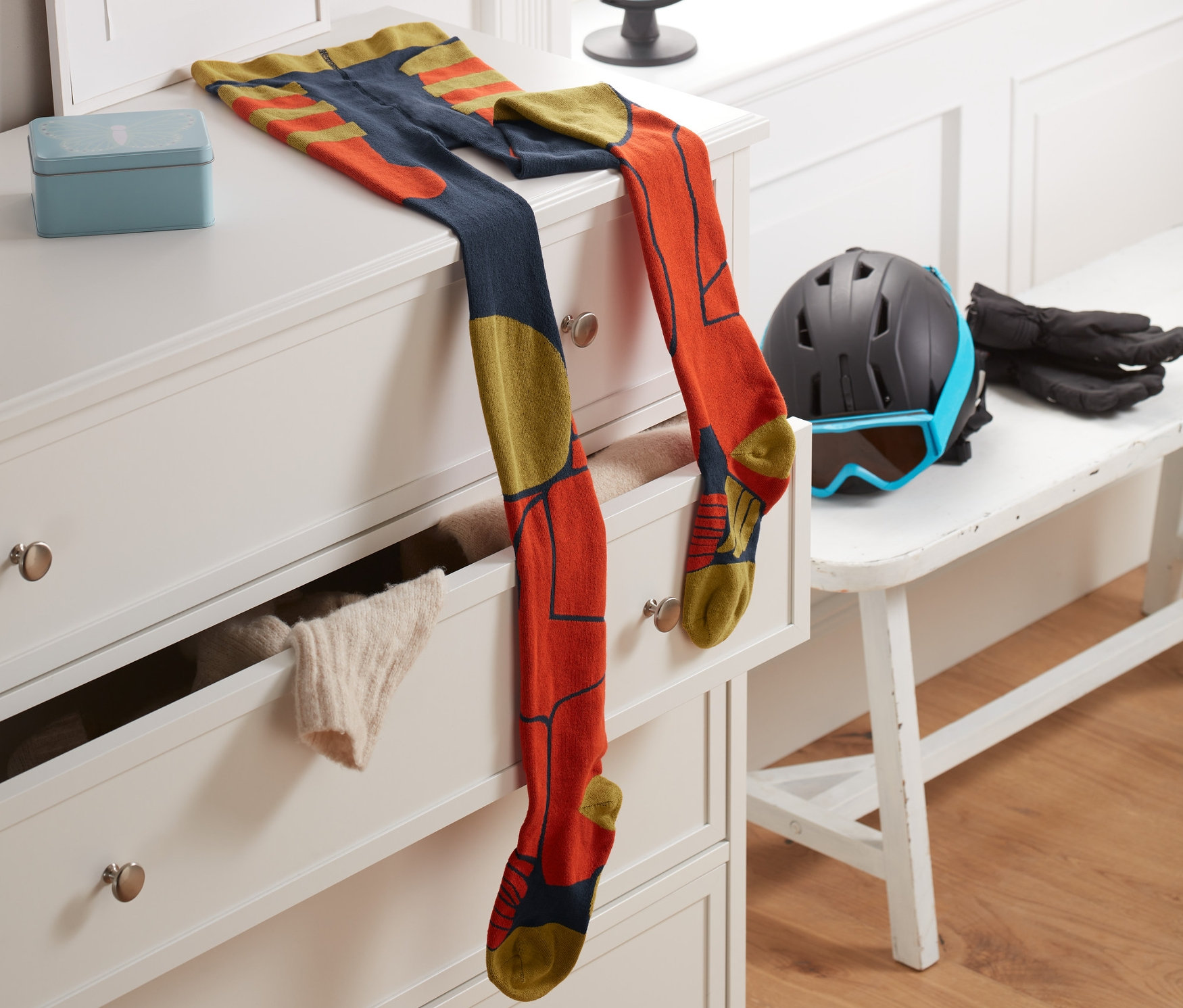 Eine Skistrumpfhose liegt auf einer weißen Kommode neben einem Rahmen mit dem Buchstaben T. Im Hintergrund befinden sich ein Fenster und ein Globus.