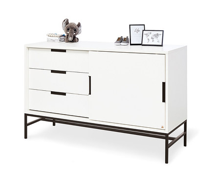 Weisse Kommode mit drei Schubladen, Schiebetüren und schwarzen Füßen. Auf der Kommode liegen ein Plüsch-Elefant, Kleidung, Babyschuhe und zwei Bilderrahmen. Zu sehen ist die Pinolino Wickelkommode »Steel«, extrabreit.