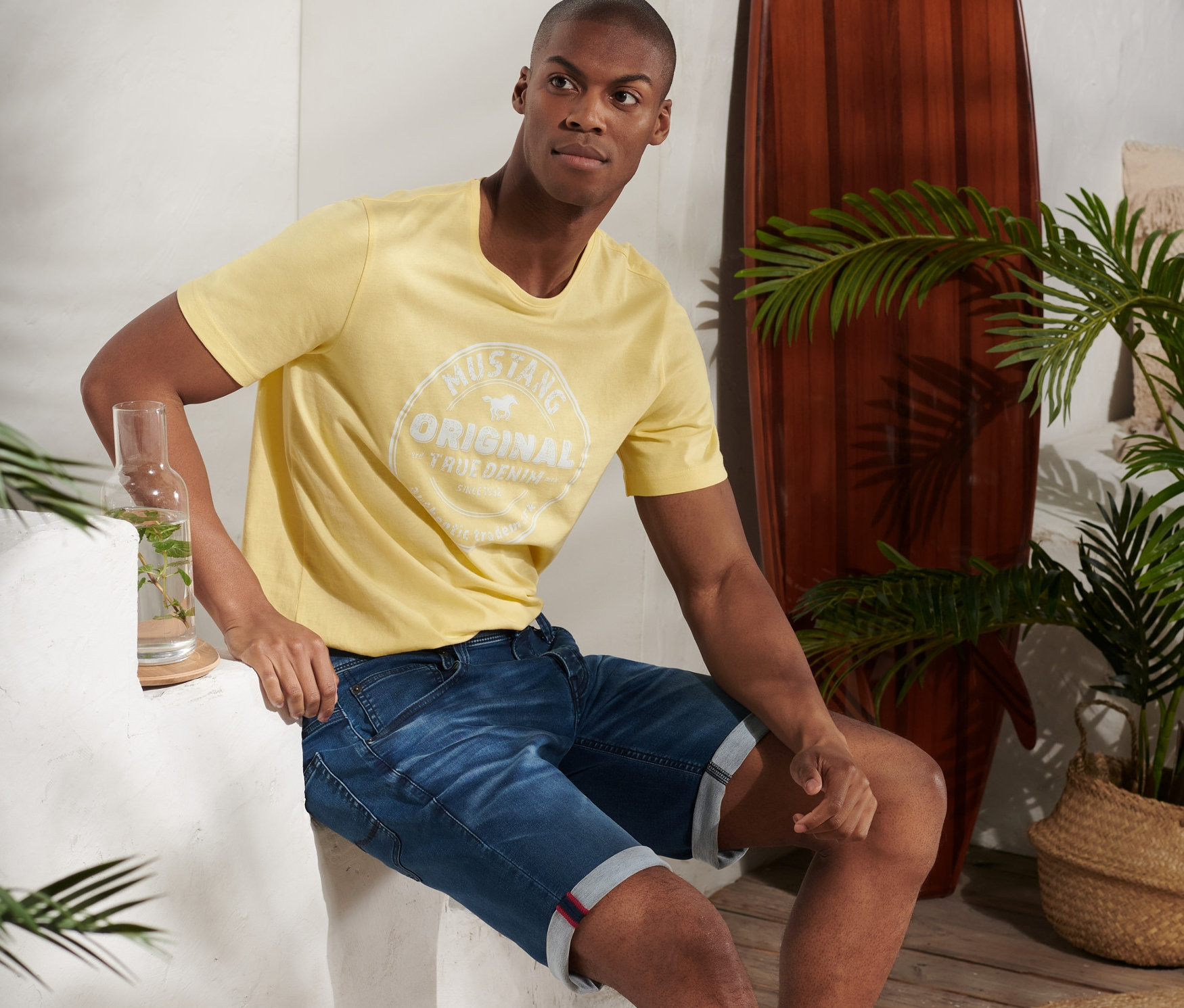 Mann sitzt und trägt blaue Jeans-Shorts »Mustang« und ein gelbes T-Shirt.
