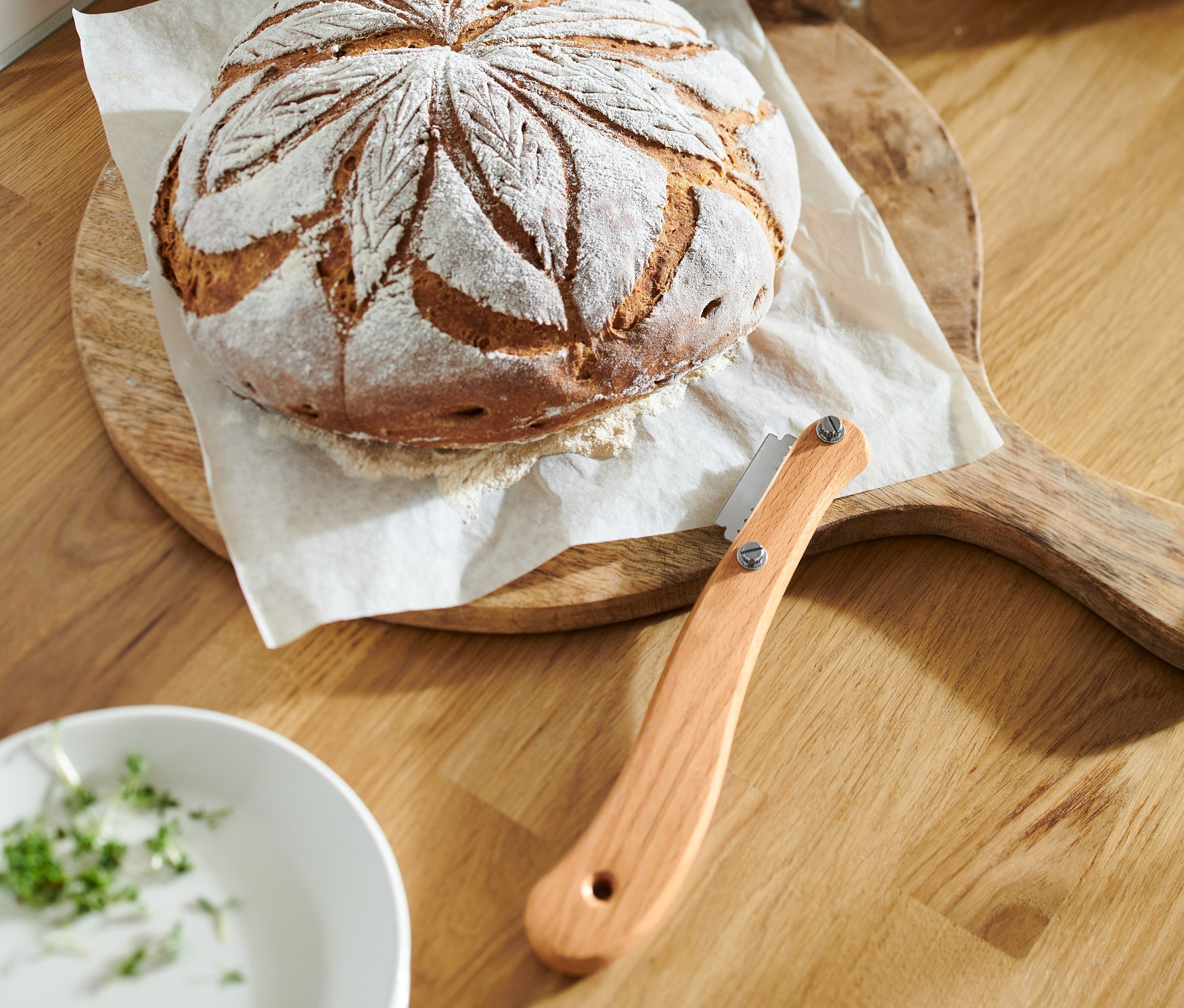 Brotlaib mit dekorativem Muster auf einem Holzbrett neben einem Bäckermesser.