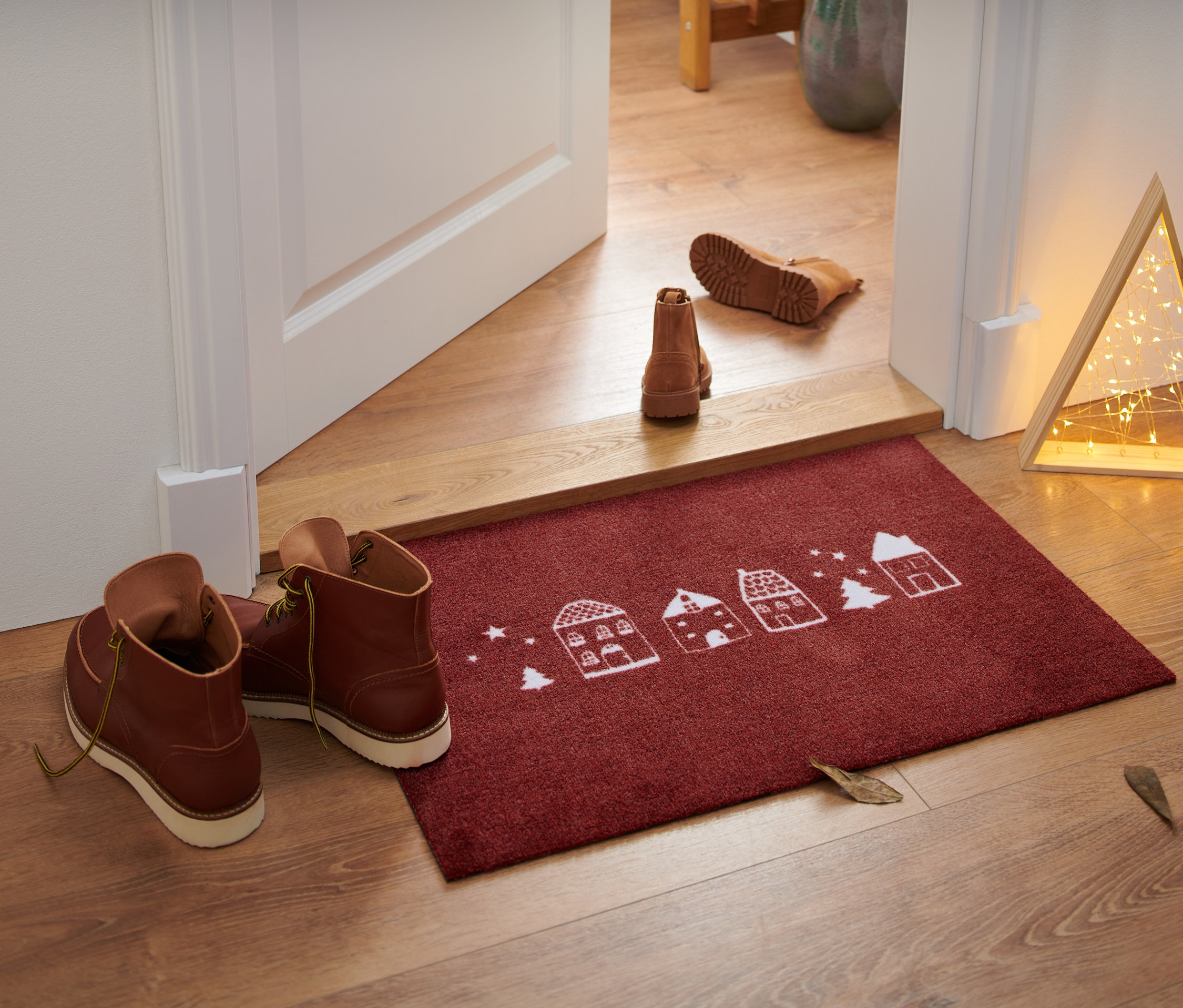 Eine rote Textil-Fußmatte mit weihnachtlichem Häuser- und Tannenbaum-Motiv liegt vor einer offenen Tür, daneben stehen braune Schuhe.