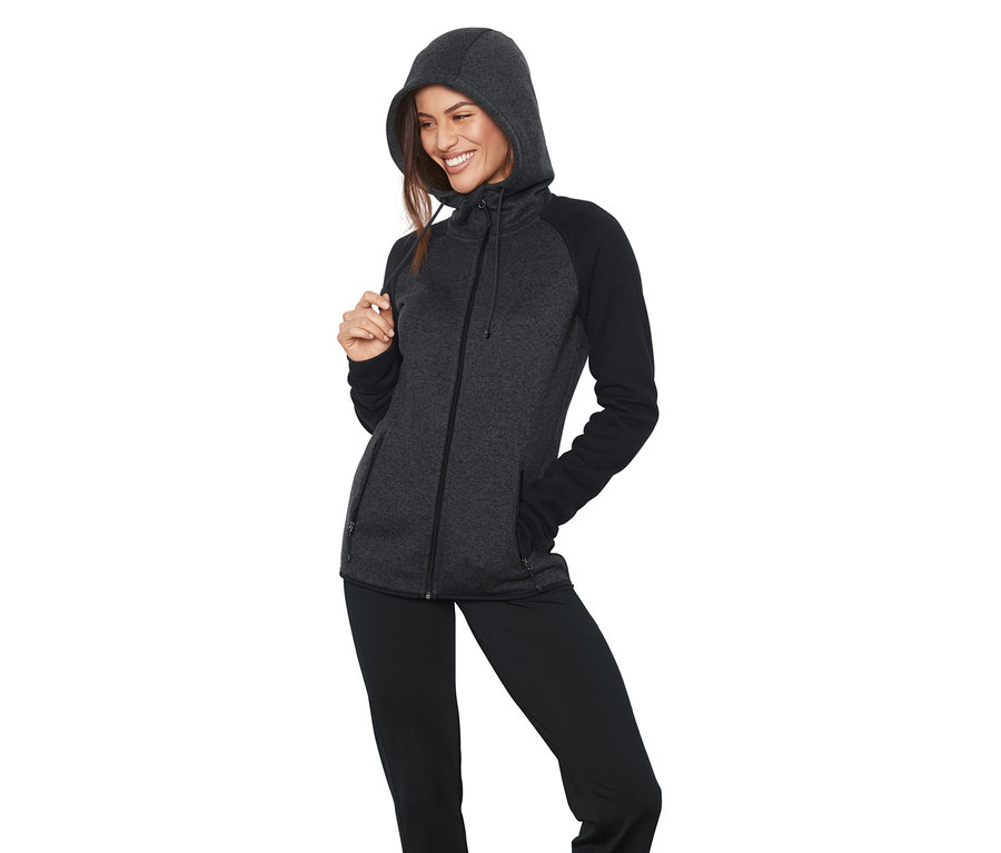 Frau posiert in einer Strickfleece-Jacke mit Kapuze und Reißverschluss. Die Jacke ist grau mit schwarzen Ärmeln.