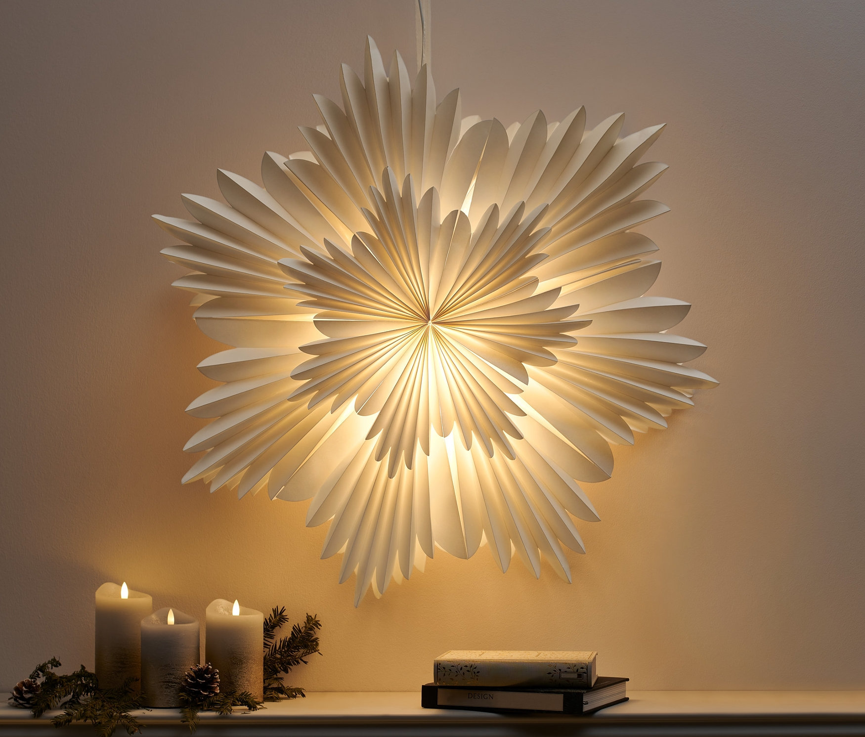 Ein hängender LED-Papierstern, XL, leuchtet über drei Kerzen und zwei Bücher auf einem Sims.