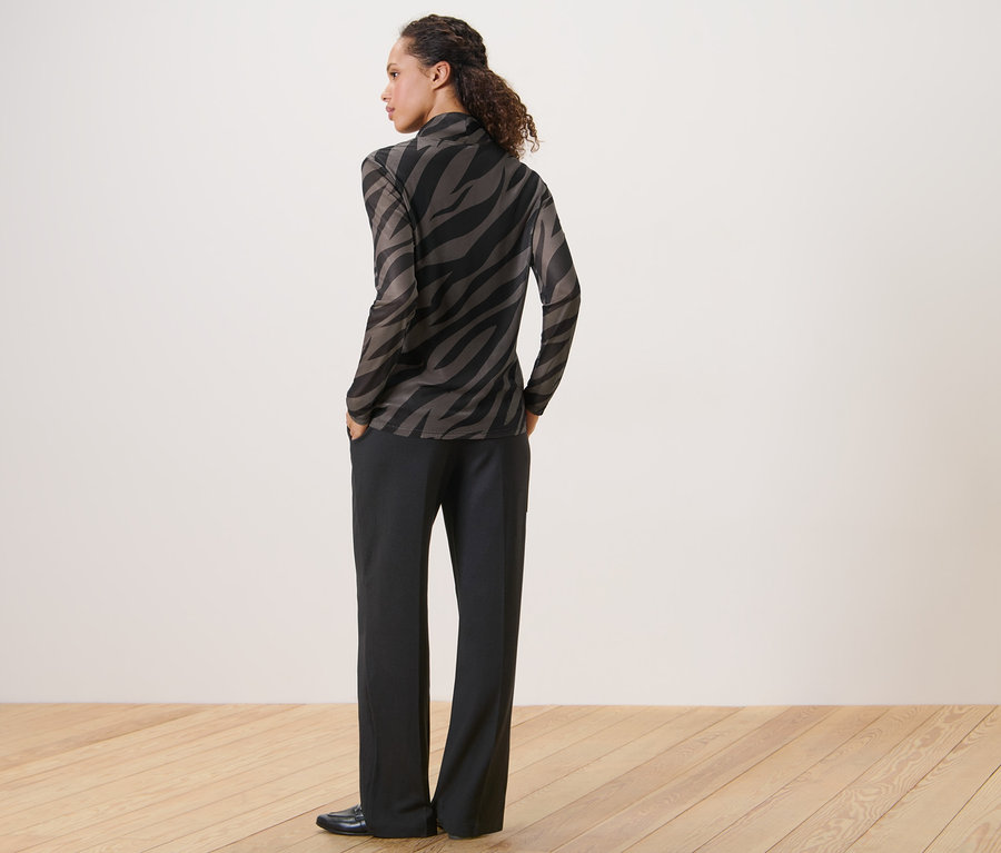 Frau posiert mit dem Rücken zur Kamera in einem bedruckten Mesh-Rollkragenshirt und einer schwarzen Hose.