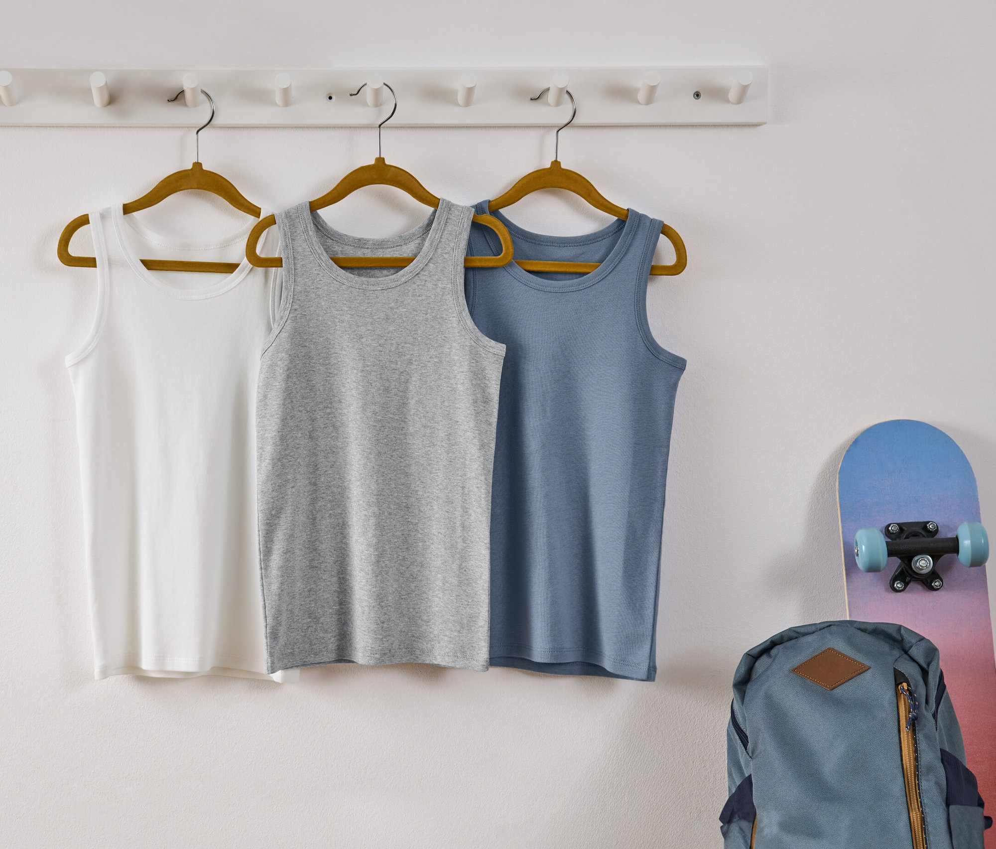 Drei Kinder-Unterhemden hängen auf Kleiderbügeln an einer weißen Wand. Die Hemden sind weiß, grau und blau.