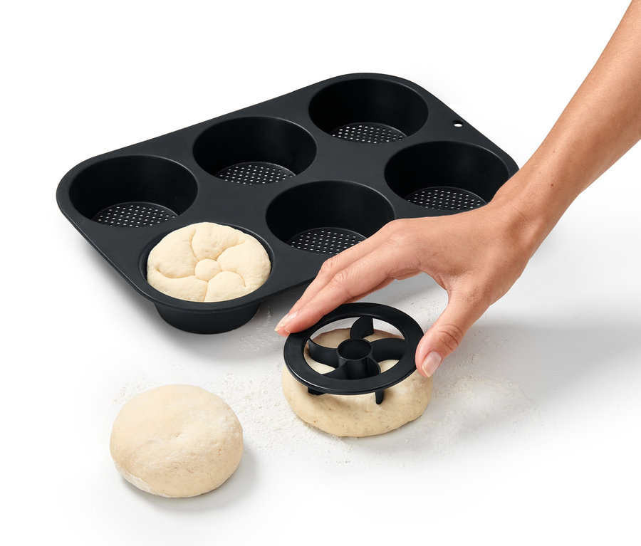 Eine Hand formt Teig für Brötchen mit einem Ausstecher neben einer perforierten Brötchenbackform.