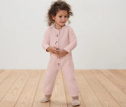 Ein kleines Mädchen steht auf einem Holzboden und trägt einen rosa Baby-Strickoverall.