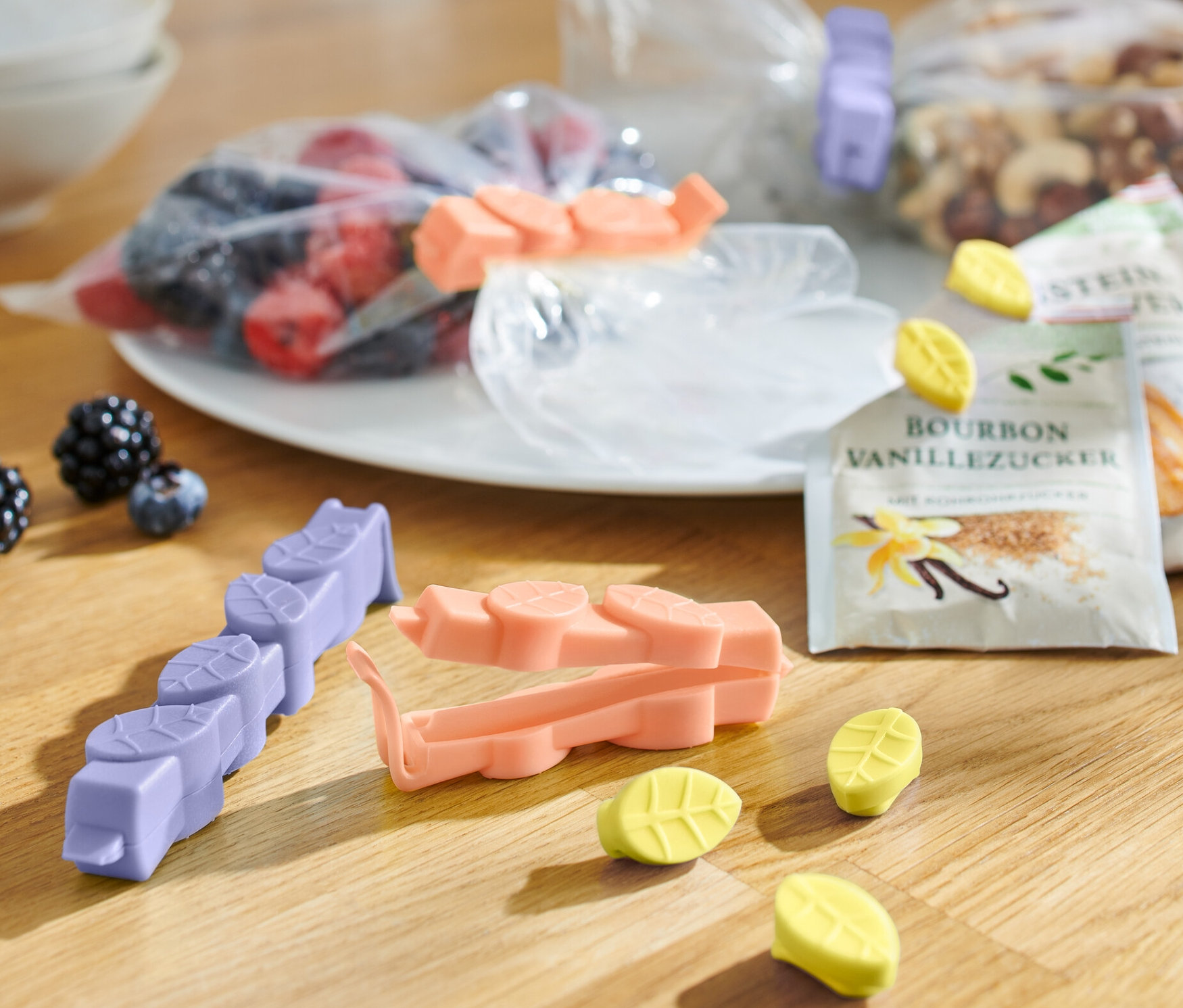 Auf einem Holztisch liegen 15 Aromaclips in verschiedenen Farben, Beutel mit Obst, Nüssen und Vanillezucker.