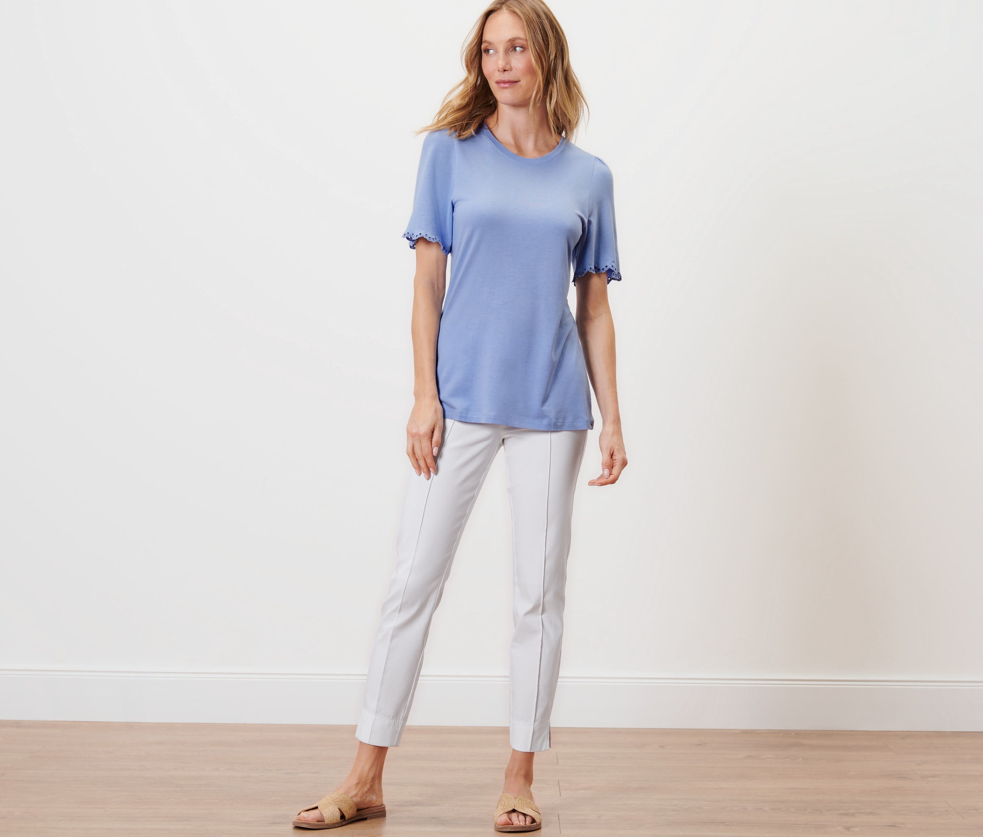 Frau steht und trägt ein hellblaues Shirt mit Raffung und eine weiße Hose.