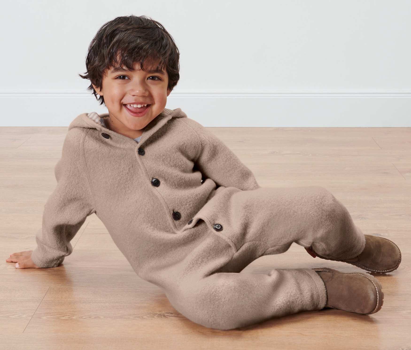 Ein Junge liegt auf dem Boden und trägt einen braunen Baby-Walkoverall mit Merinowolle.