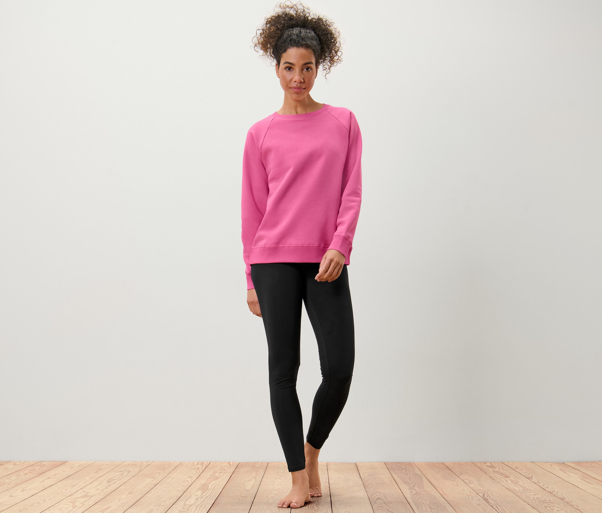 Frau steht barfuß und trägt ein Yogasweatshirt in Pink und schwarze Leggings.
