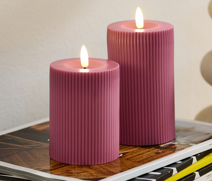 Zwei rosafarbene LED-Echtwachskerzen stehen auf einem Stapel Zeitschriften. Im Hintergrund eine Vase mit Eukalyptus.
