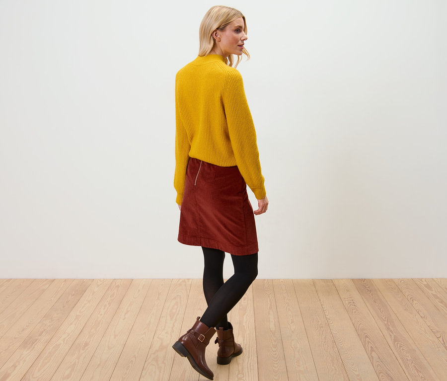 Blonde Frau trägt gelben Pullover und Cordrock. Sie steht auf einem Holzboden.