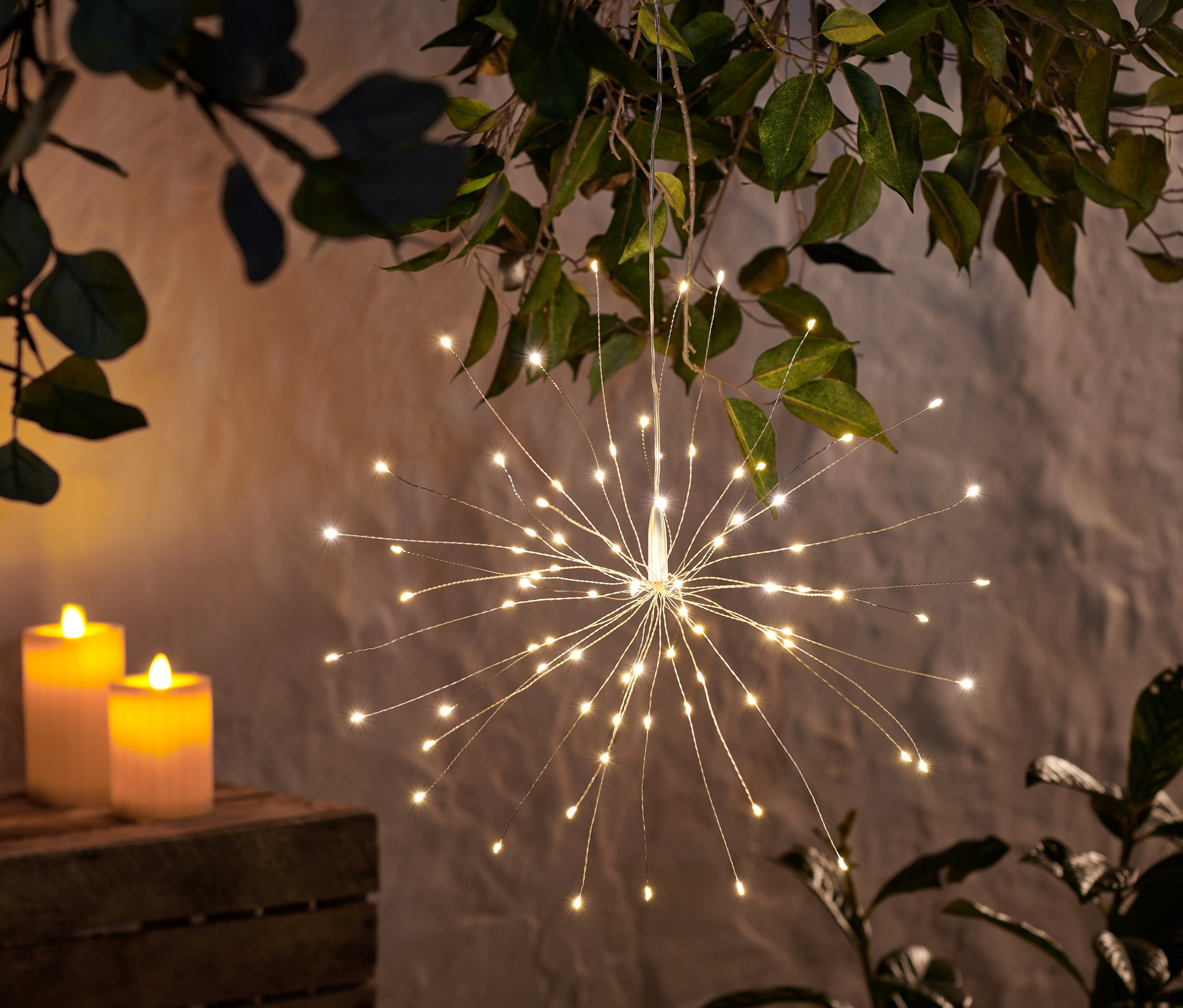 Ein LED-Leuchtzweig hängt von einem Baum, daneben stehen zwei zylindrische Kerzen und ein Blumentopf.