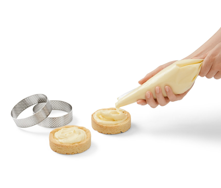 Eine Frau verziert zwei Törtchen mit Creme. Daneben liegen 4 Edelstahl-Backringe.