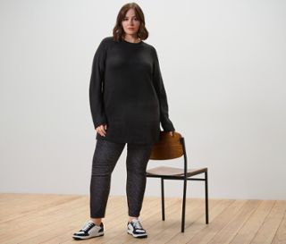 Frau posiert in langem schwarzen Strickpullover und bedruckter Bengalin-Hose neben einem Stuhl.