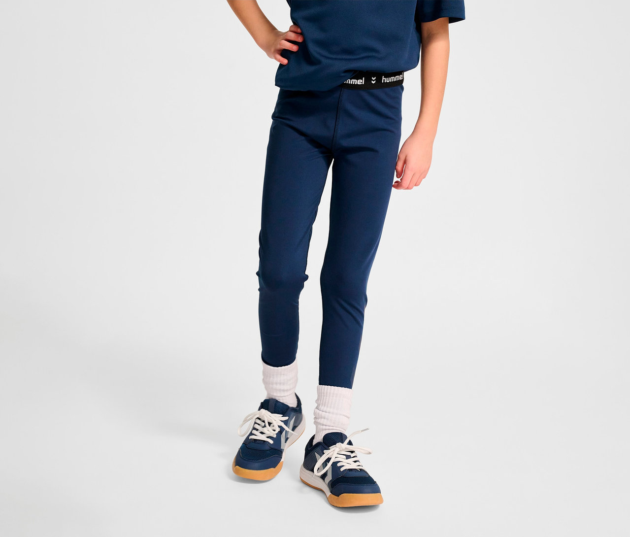 Kind steht vor einem weißen Hintergrund und trägt blaue HUMMEL HMLJR Pulse Mid Waist Tight, ein blaues T-Shirt und Turnschuhe.