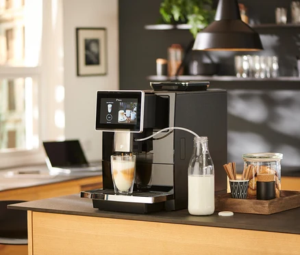 Kaffeevollautomat bereitet Latte Macchiato in einem Glas zu. Daneben stehen eine Milchflasche und ein Glas mit Keksen.