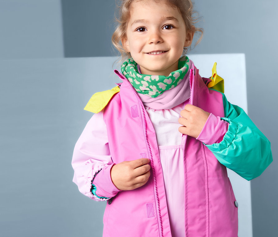 Mädchen schliesst eine pinke Jacke und trägt ein recyceltes Multifunktionstuch, rosa-grün.