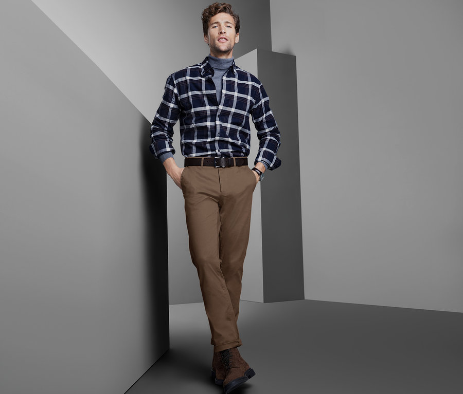 Mann posiert an einer Wand in einem karierten Hemd, braunen Chinos und braunen Schuhen.