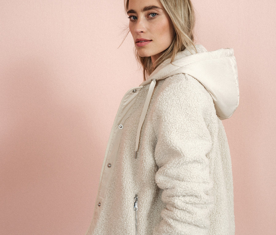 Frau blickt über die Schulter und trägt einen cremefarbenen Mantel mit Kapuze.