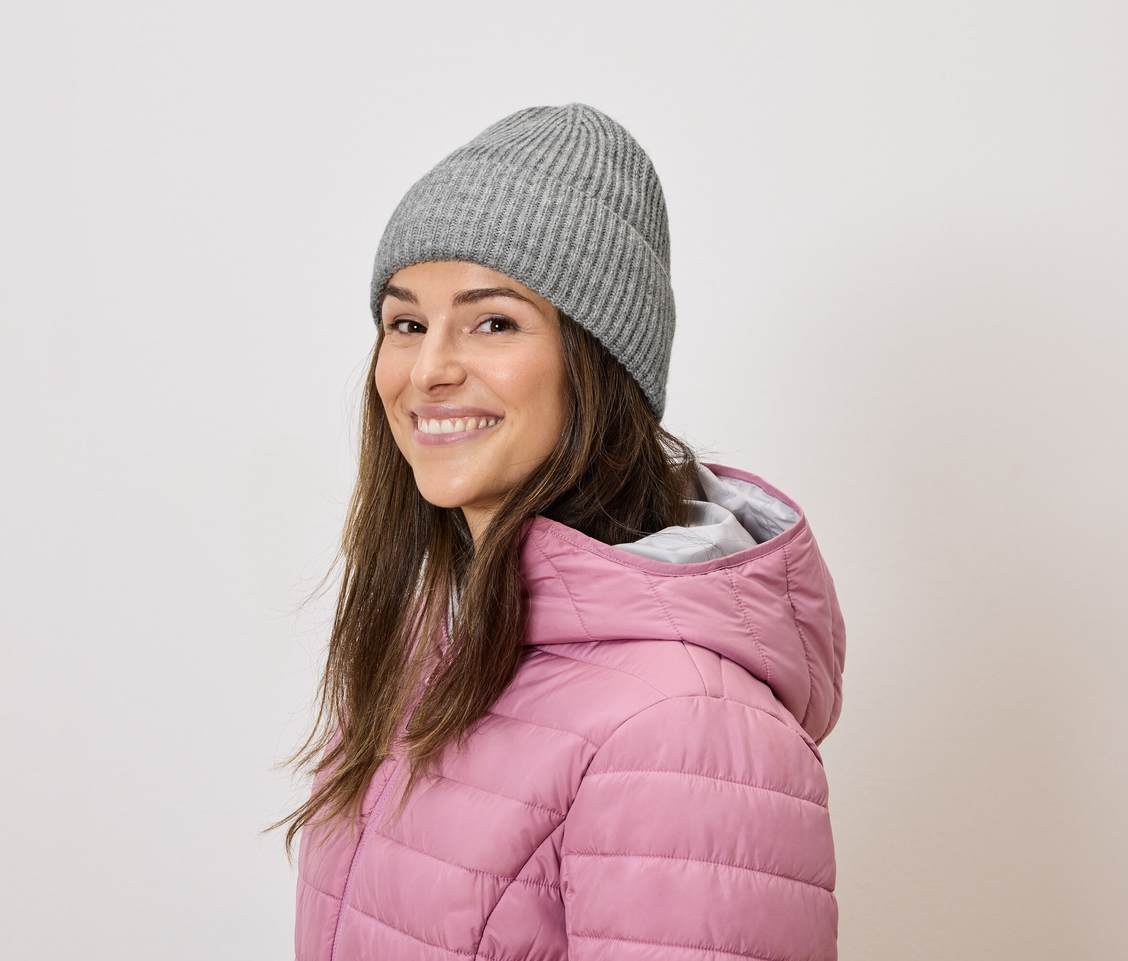 Frau mit braunen Haaren lächelt und trägt eine graue Strickmütze und eine rosa Daunenjacke.