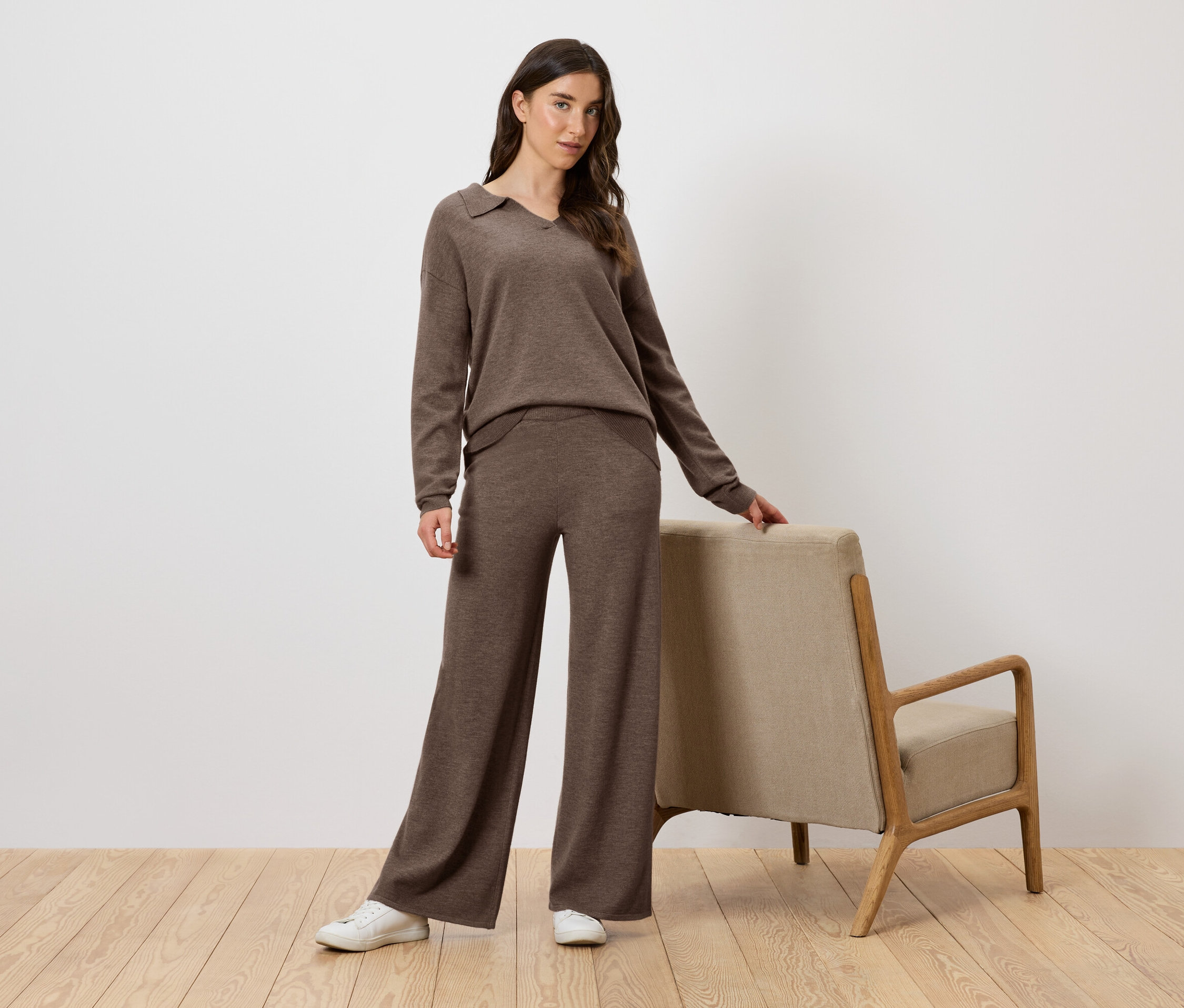 Frau im braunen Strickset posiert neben einem Sessel.