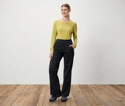 Eine Frau steht auf einem Holzboden. Sie trägt eine grüne Bluse mit langen Ärmeln und eine schwarze Hose.