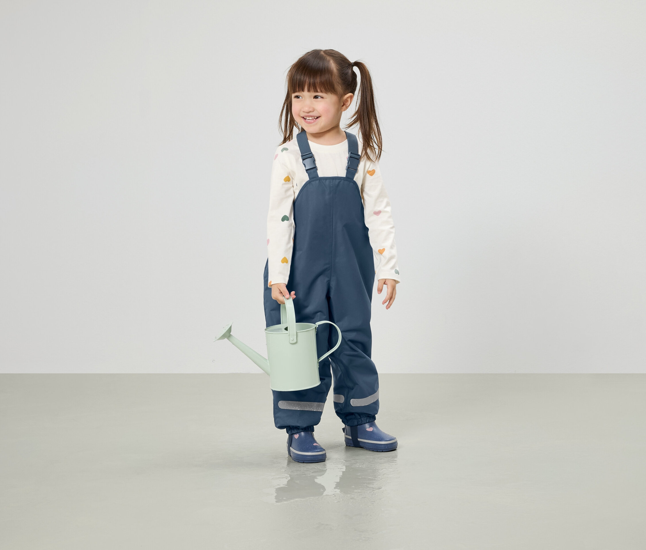 Lächelndes Mädchen mit Zöpfen hält eine hellgrüne Gießkanne, gekleidet in blaue Hosen und Gummistiefel.