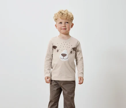 Ein Junge mit blond gelocktem Haar trägt ein beiges Langarmshirt mit braunen Bärengesicht-Applikationen und braune Hosen.
