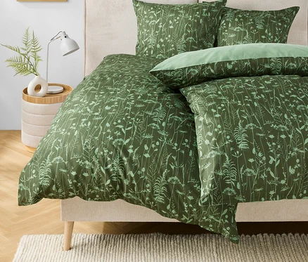 Grüne Bettwäsche mit Blumenmuster auf einem Bett. Auf dem Nachttisch stehen eine Lampe und eine Vase mit einer Pflanze.