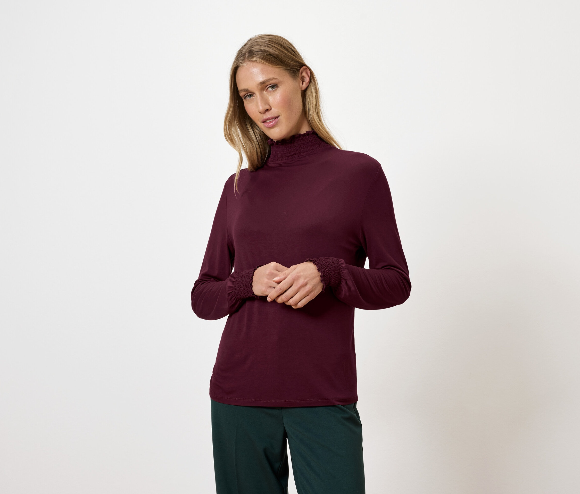 Blonde Frau trägt ein weinrotes Rollkragenshirt und grüne Hosen.