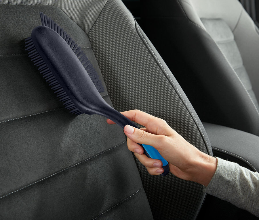 Eine Hand hält eine Bürste mit blauen Akzenten und reinigt einen grauen Autositz.