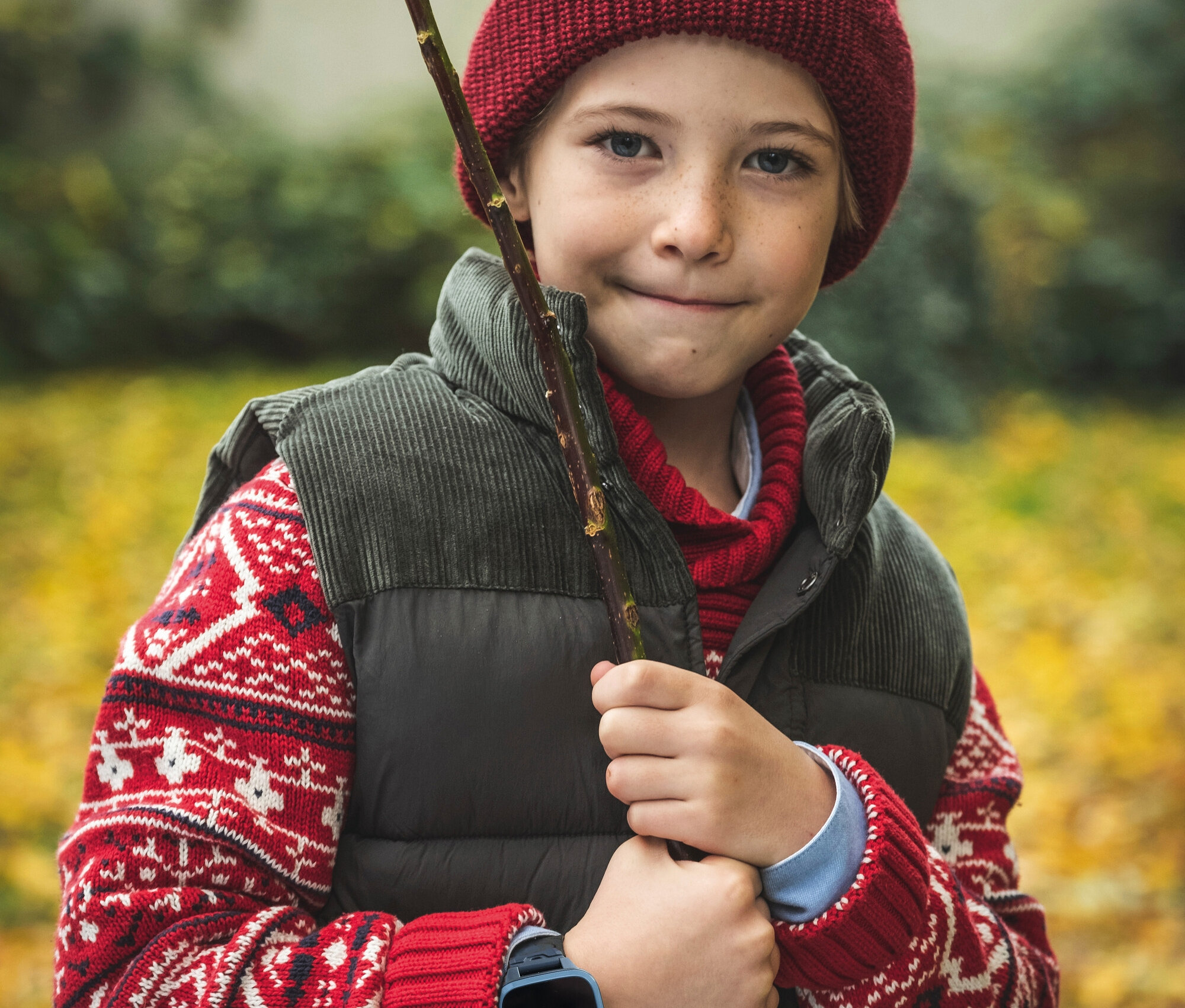 Junge mit roter Mütze, Pullover und Weste hält einen Stock. Er trägt eine schwarze Kinderuhr am Handgelenk.