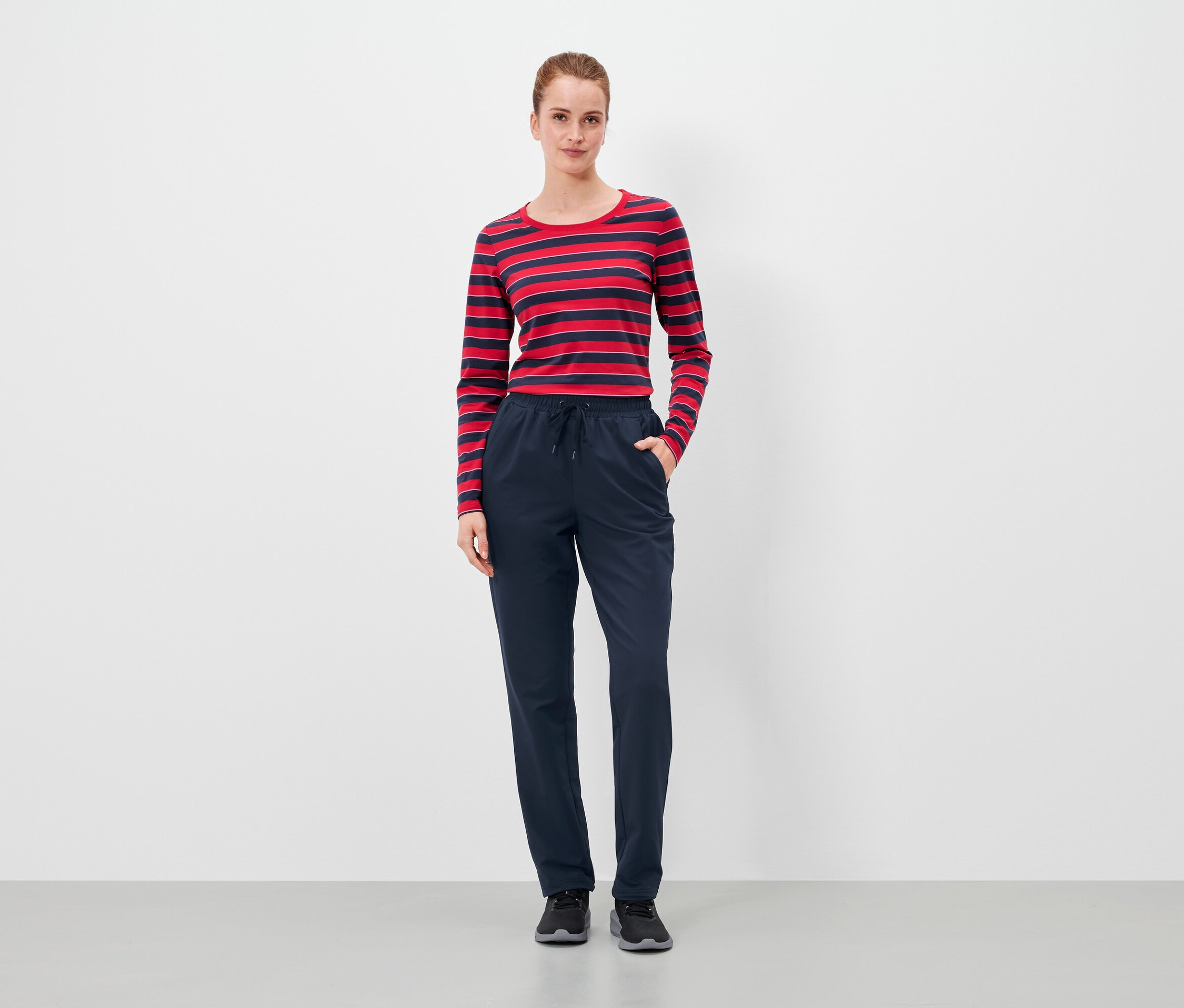 Frau steht mit Händen in den Taschen einer dunkelblauen Hose und trägt ein rot-blau gestreiftes Langarmshirt.