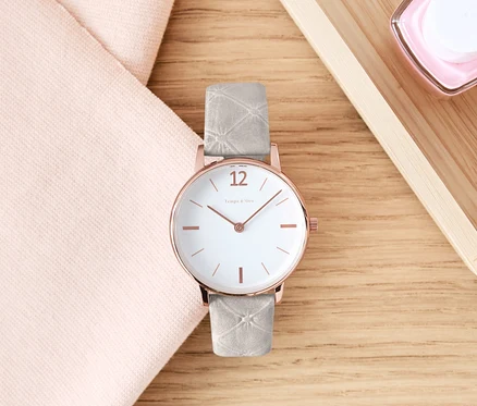 Eine Armbanduhr mit Lederband liegt auf einem Holztisch neben einem rosa Tuch und Kosmetika.