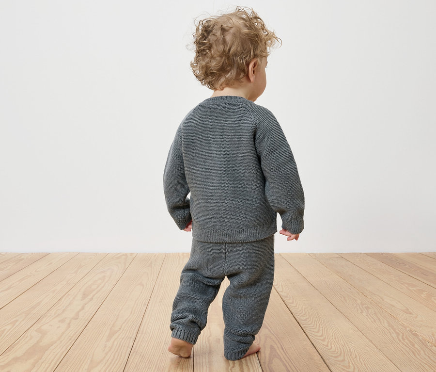 Ein Kind mit lockigen Haaren trägt von hinten eine Baby-Strickkombi.