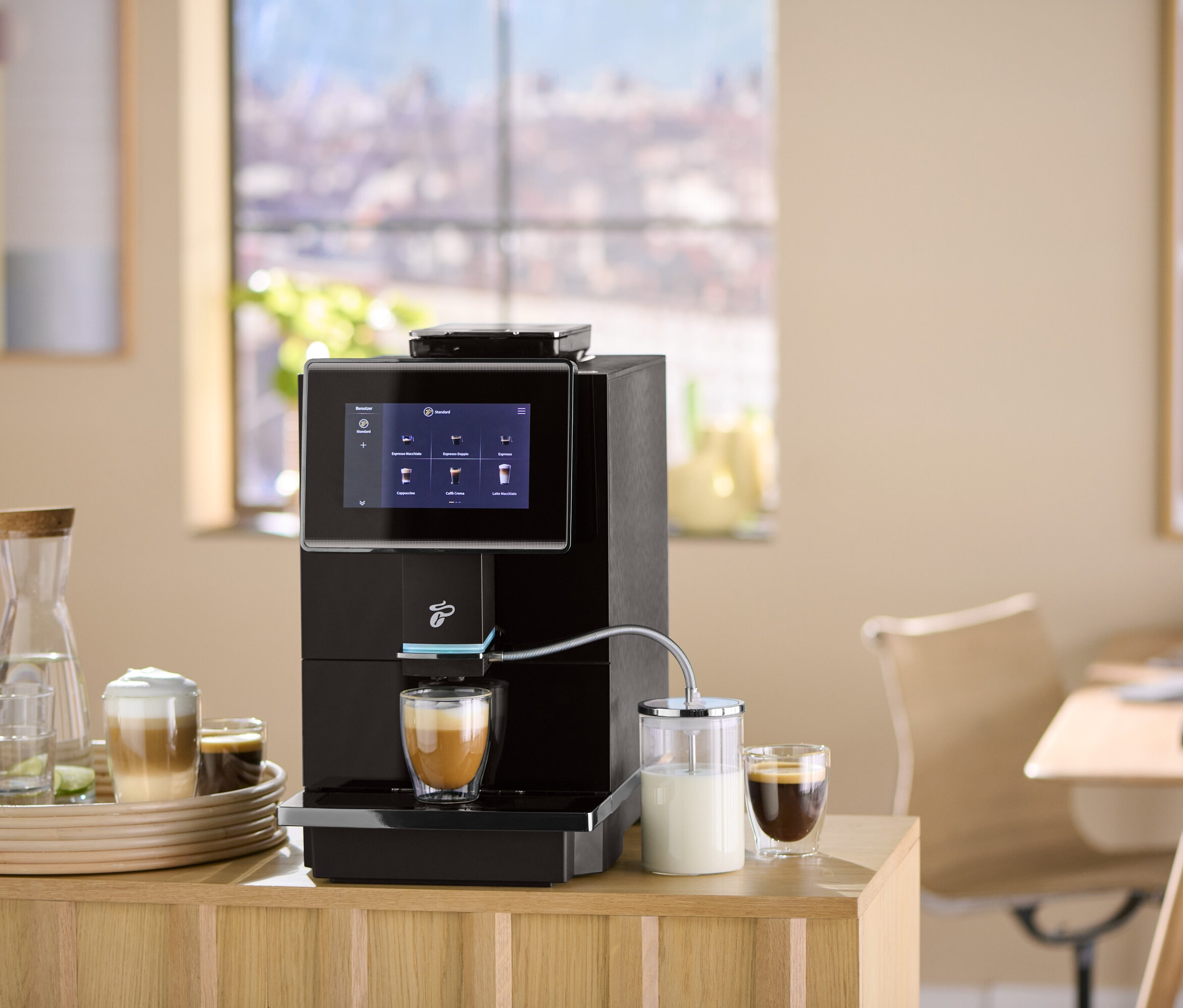Ein schwarzer Kaffeevollautomat bereitet Kaffee in ein Glas zu. Auf einem Tablett befinden sich Gläser, eine Karaffe und ein Kaffee mit Schaum.
