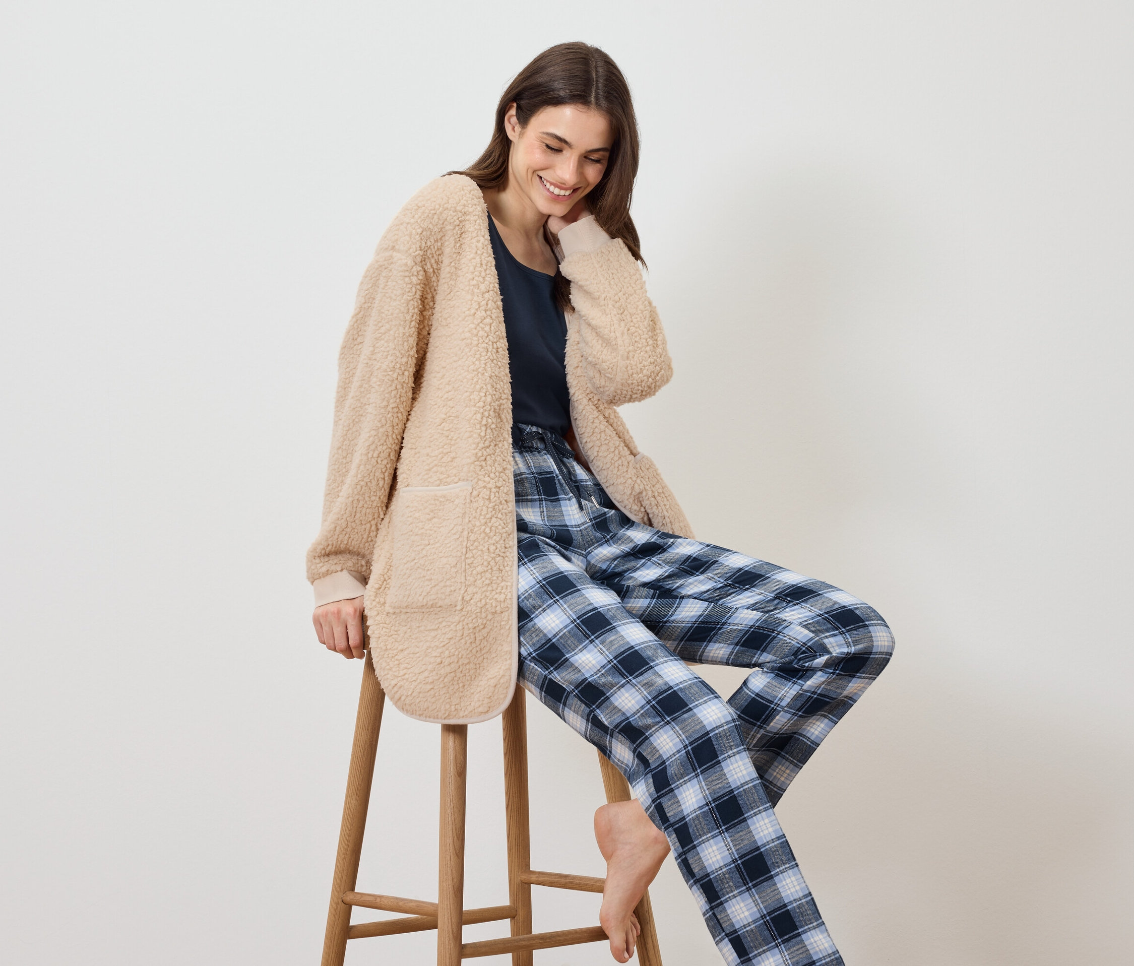 Frau sitzt auf einem Hocker, trägt eine beige, flauschige Jacke und eine blaue, karierte Pyjamahose.
